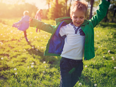 Child running through a sunlit grassy field with arms outstretched, while other children play in the background. Warm sunlight and green outdoor surroundings create a bright spring scene.