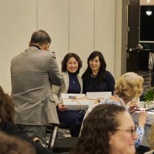 Two females sitting at table, smiling and posing for a picture. Male is standing and taking the photo.