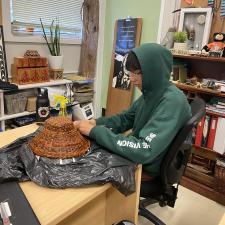 Student sitting at desk working on cedar weaving at hat