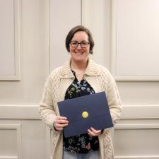 Female ABC award recipient stands in front of wall holding certificate