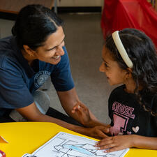 Female parent smiles at female student while sitting at a school table looking at school work