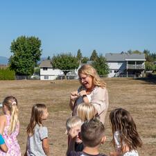 Female principal talks with a group of young elementary students with the school field in the background.