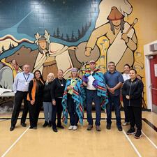 Group of people standing in a gymnasium with Indigenous mural on wall behind them. Two of the individuals have been blanketed as an honouring.